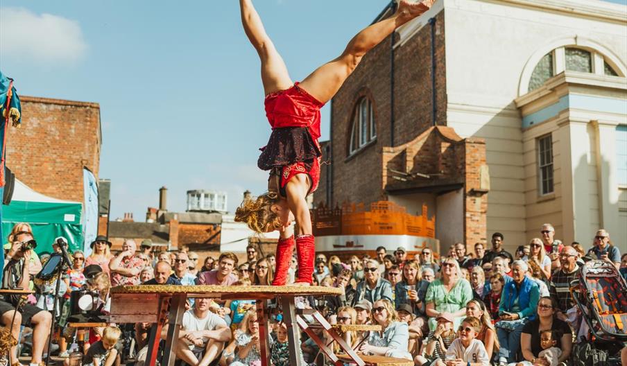 A performer doing a hand stand on a pedestal, surrounded by a crowd.