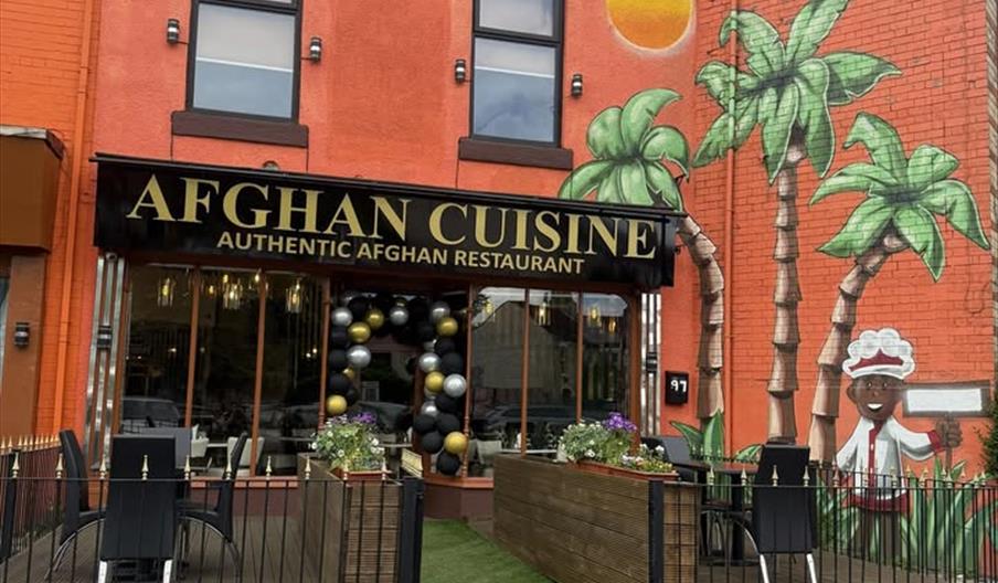 The image shows the exterior of a brightly colored Afghan restaurant with a sign reading "Afghan Cuisine Authentic Afghan Restaurant." The front patio
