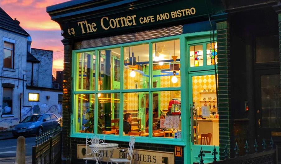 A brightly lit café called "The Corner Café and Bistro" with large green-framed windows and a small white table with two chairs outside, set on a stre