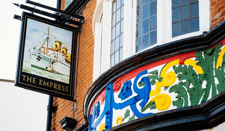 A hanging sign for "The Empress" pub featuring an illustration of a large ocean liner is mounted on a brick building with colorful, ornate painted tri