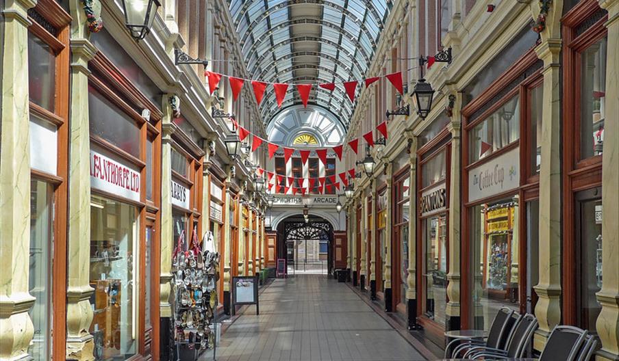 A view down Hepworth Arcade, a glass-roofed victorian shopping arcade with assorted shops down either side and red bunting hanging across the way. Vic