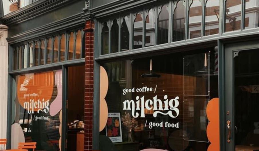 Outdoor view of "Milchig" Cafe, with large windows featuring text that reads "good coffee / milchig / good food," and several orange tables and chairs