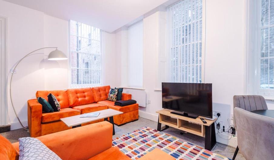 A bright living room with orange sofas, a modern floor lamp, a colorful patterned rug, a flat-screen TV on a wooden stand, and large windows with vert