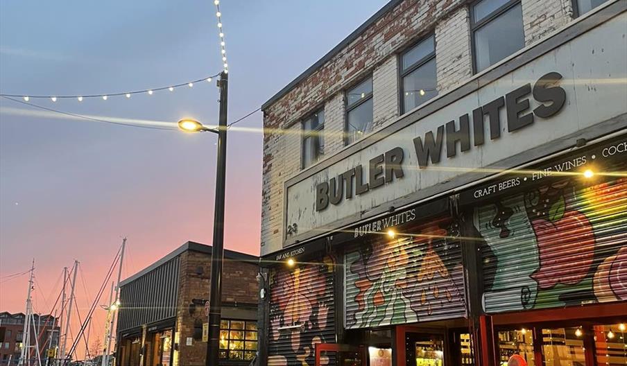 Exterior of restaurant with in an evening, with illuminated sign, roped off street level terrace, mutiple tables and chairs.