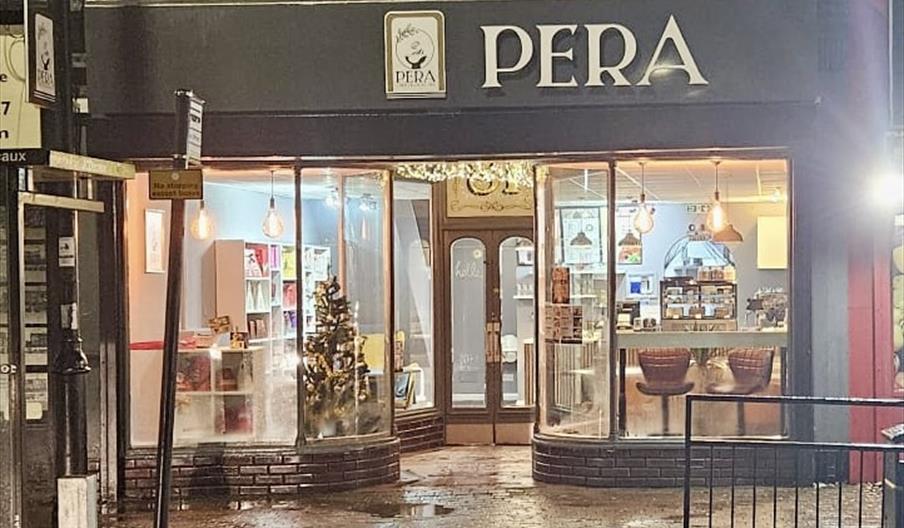 Exterior view of PERA Cafe at night, with large front windows showing a lit-up interior featuring shelves, a counter, and a decorated Christmas tree.