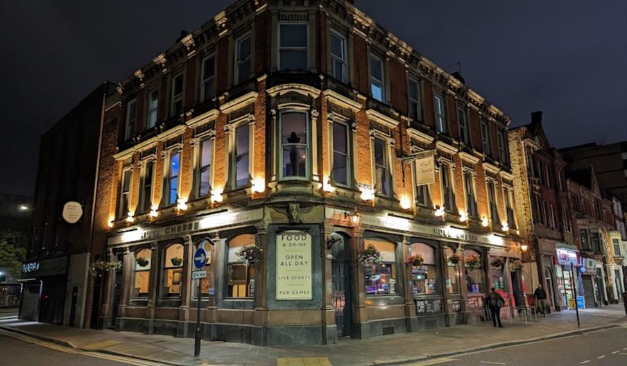 Exterior of Hull Cheese. A large, ornate brick pub building with illuminated windows and exterior lights stands on a street corner at night, with a si