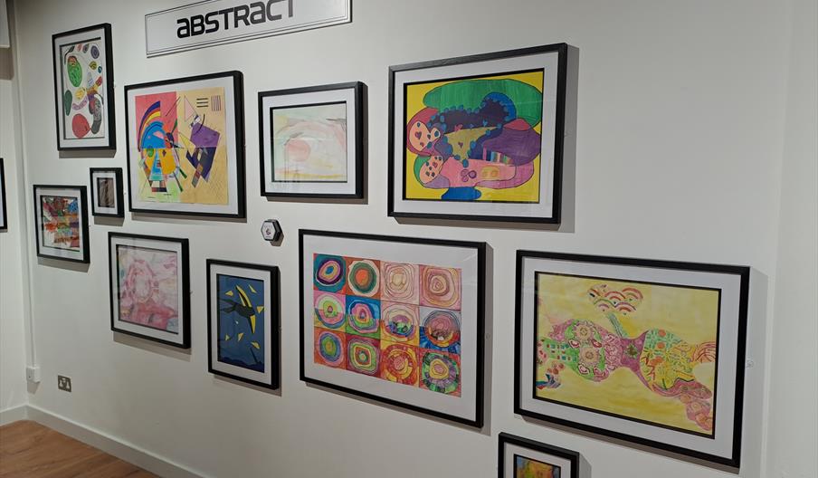 A gallery wall displays a collection of framed colorful abstract artworks, arranged in a grid and labeled with a sign that says "ABSTRACT." The drawin