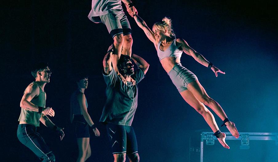 Four acrobats perform on stage, with one man lifting another by the legs above his head and a woman in midair, supported by holding hands with the man