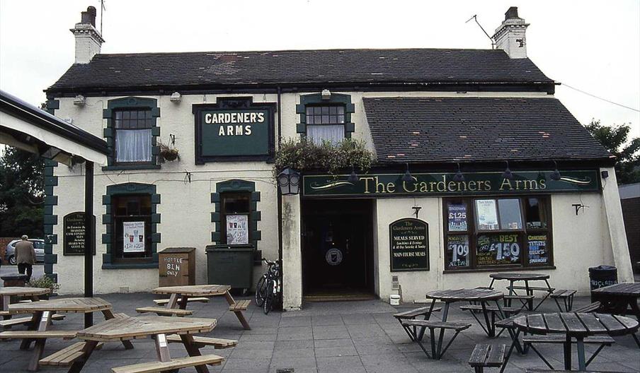 A traditional English pub called "The Gardeners Arms" with a white exterior, green trim, and multiple windows is shown. Several wooden picnic tables a