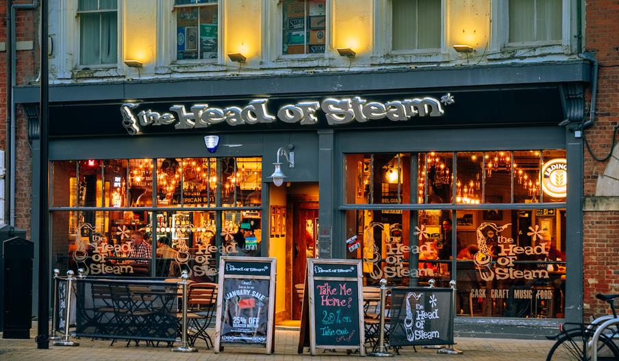 Exterior of  pub called "The Head of Steam" with illuminated signage, large windows with hanging string lights, outdoor seating and chalkboard signs a