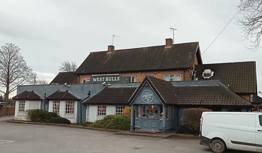 A close-up of a cheeseburger with lettuce, tomato, onions, and bacon on a sesame seed bun, served with a pile of French fries on a plate. In the backg