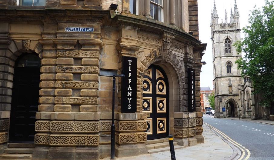 A street corner with an ornate stone building featuring a black and gold door and two vertical signs reading "TIFFANYS." The street is named Bowlalley
