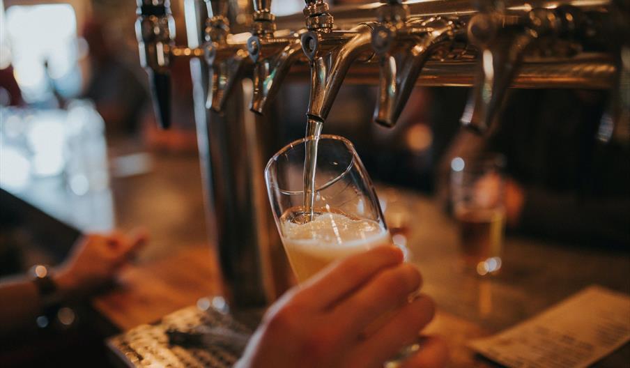 A person is holding a tilted glass under a beer tap, filling it with light-colored beer at a bar with several other taps in the background.