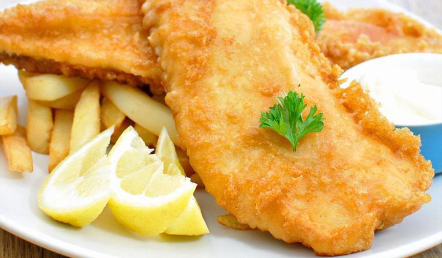 A plate of golden fried fish fillets served on top of thick-cut French fries, garnished with fresh parsley, with lemon wedges and a small container of