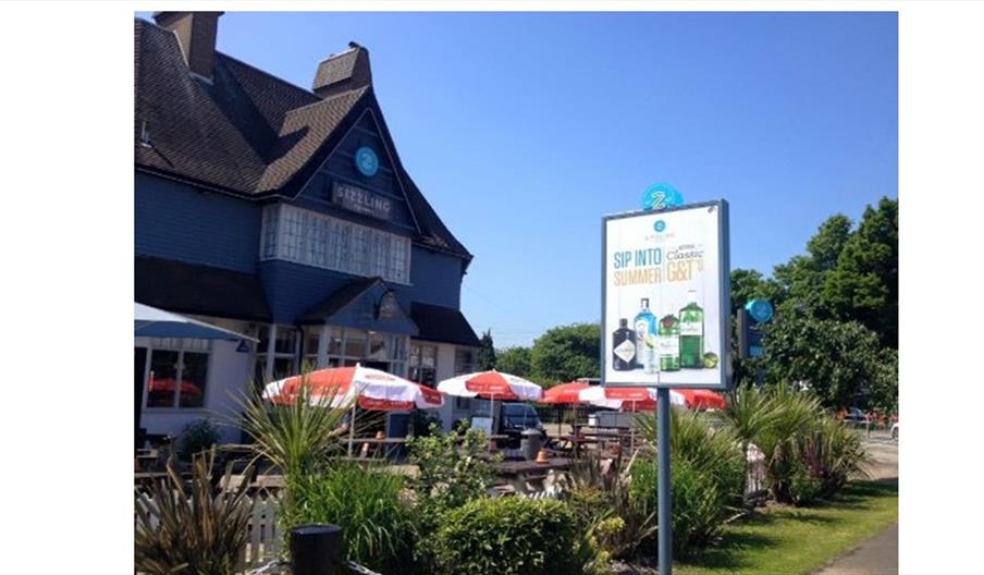Outdoor seating area of The Hop Pole Hotel with red-and-white umbrellas, green plants, and a large roadside sign advertising summer gin and tonic drin