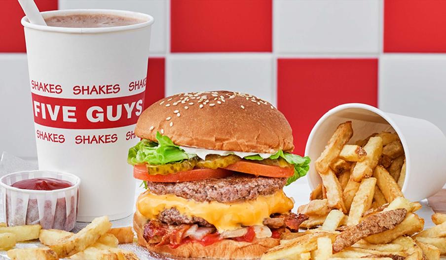 A Five Guys meal featuring a double cheeseburger with lettuce, tomato, and bacon, accompanied by a cup of fries spilling onto the table, a ketchup con