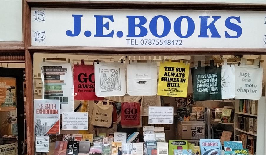 Front window of J.E. Books bookshop, displaying books, event posters, and reading-themed tote bags hanging above the entrance.
