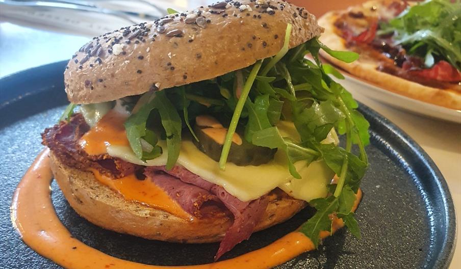 Close-up of a gourmet bagel served at Heavenly Desserts Hull, filled with sliced pastrami-style beef, melted cheese, crispy bacon, pickles and fresh r
