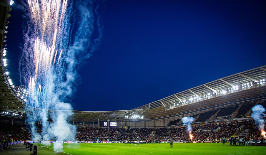 Fireworks and smoke rise into a deep blue evening sky inside a floodlit MKM stadium, with a green pitch below and packed spectator stands surrounding