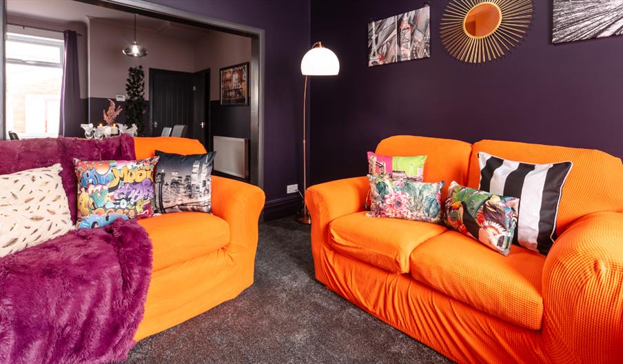Living room with bright orange sofas, colourful cushions, and deep purple walls.