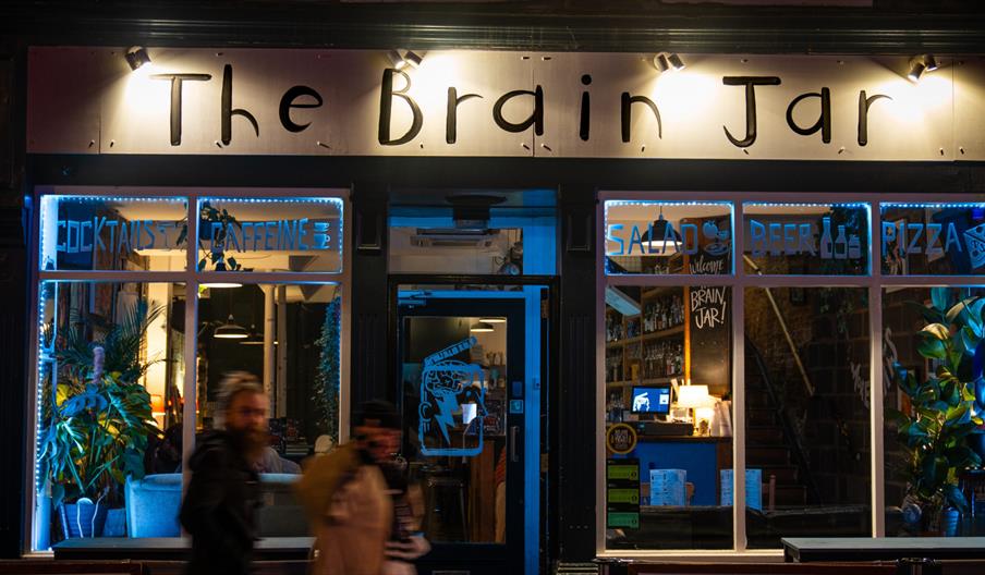 Exterior of The Brain Jar bar in Hull at night with lit signage and cosy interior visible through windows.
