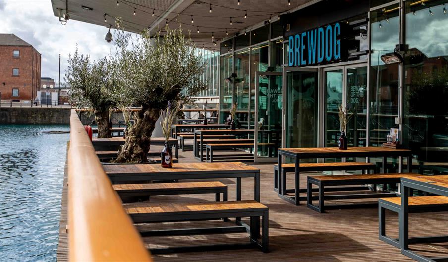 Outdoor seating area at BrewDog Hull overlooking Princes Dock, with wooden picnic benches, waterside views and the brewery's modern glass-fronted venu