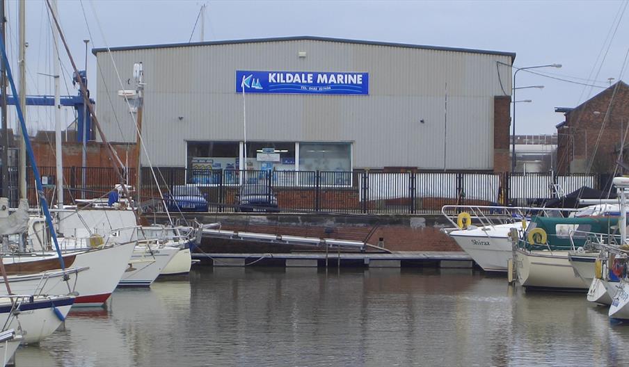 Boats moored in Hull marina with Kildale Marine building in the background, reflected in calm water on an overcast day.