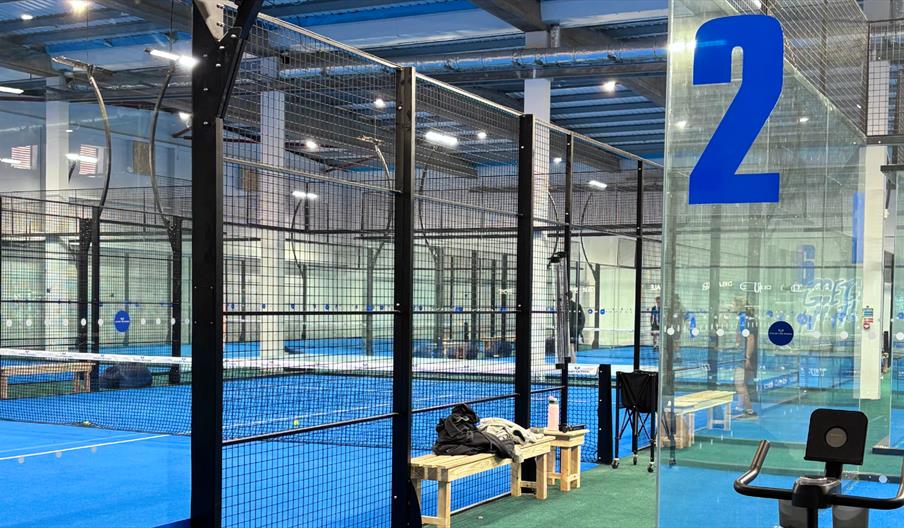 Indoor padel courts at Court De Padel Hull with blue playing surfaces, glass walls, and mesh fencing, including clearly numbered courts, seating bench