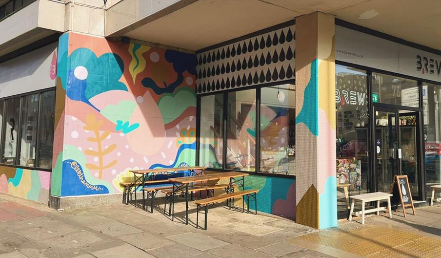 Exterior of Brew bar and café in Hull city centre, featuring a colourful mural-covered façade, outdoor picnic-style seating, and a welcoming entrance
