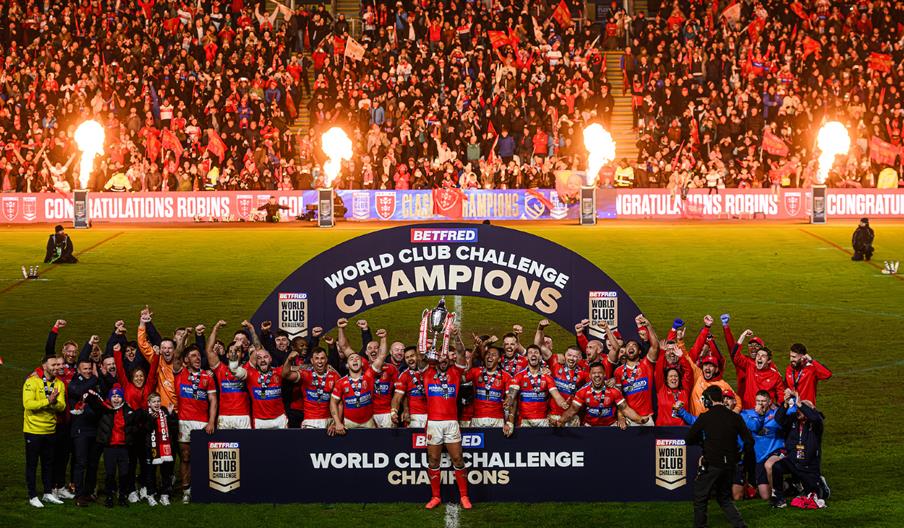 Hull KR Team in red shirts celebrate on the pitch, lifting a trophy beneath a large “Betfred World Club Challenge Champions” arch, while a packed stad