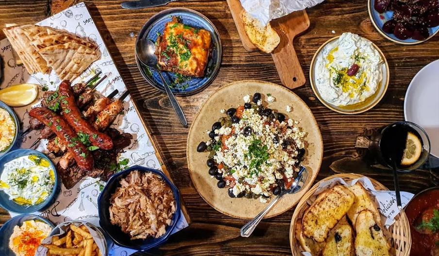 A wooden table filled with various Greek dishes, including skewered meats, pita bread, fries, tzatziki, rice, a Greek salad with olives and feta, gril