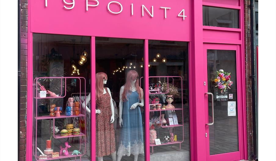 A brightly coloured storefront with bold pink framing and large glass windows displays two mannequins in dresses and several eclectic items on shelves