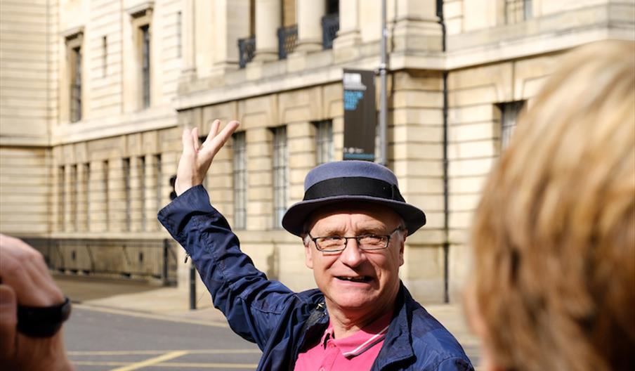 A man in a pork pie hat gestures at the building behind him animatedly.