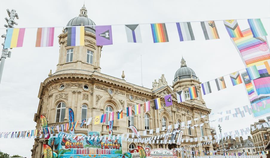 Colourful bunting with several LGBT flags flying in front of the Maritime Museum, with crowds of people walking underneath.