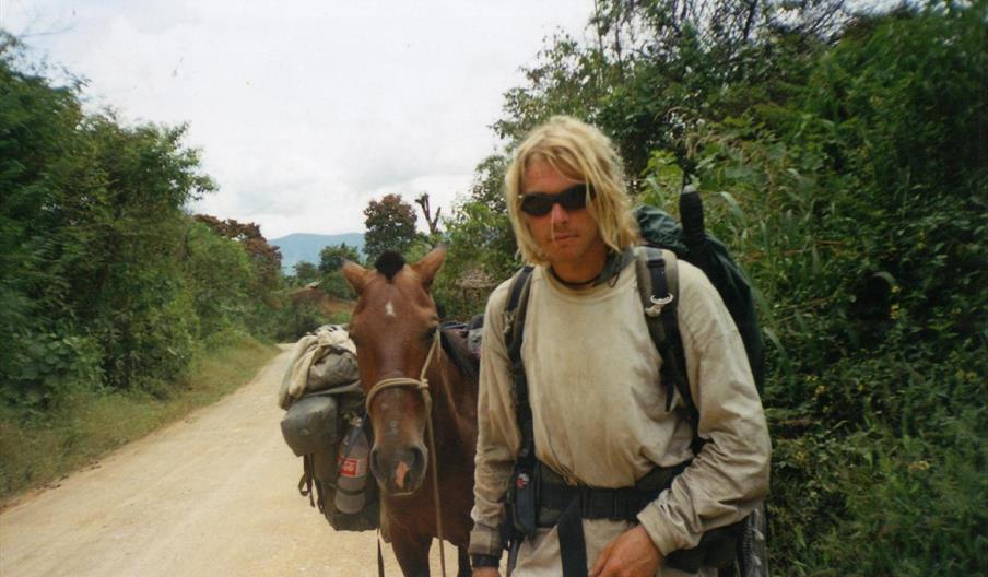 A person wearing outdoor clothing and sunglasses stands beside a packed horse on a dirt road surrounded by dense green vegetation. The person carries