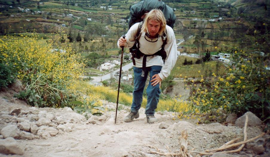 A person with long blonde hair, wearing a large backpack and using a walking stick, hikes up a rocky, dirt trail surrounded by yellow wildflowers, wit