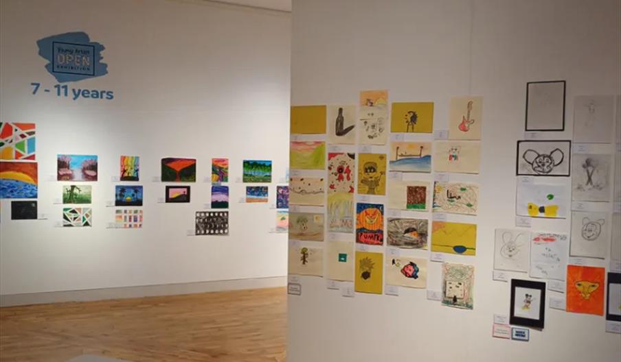 A gallery wall displays colorful artwork created by children aged 7 to 11, featuring a variety of paintings and drawings arranged in neat rows on whit