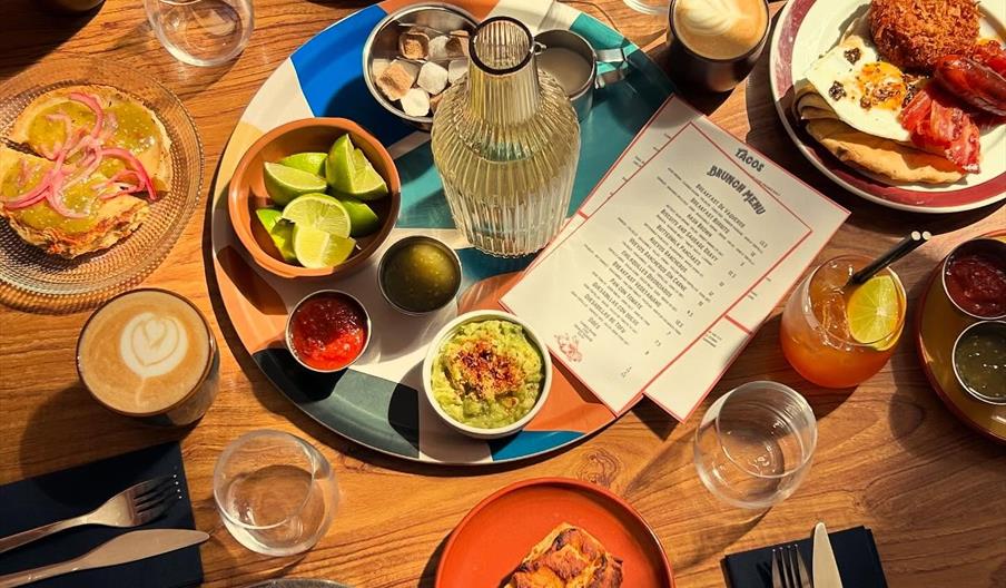A wooden table in Food For Cowboys set for brunch features assorted plates of food, including pancakes, avocado toast, pastries, eggs with bacon, and