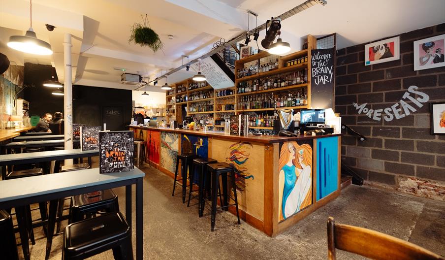 Interior of The Brain Jar. A cozy bar with wooden shelves filled with bottles behind the counter, colorful murals painted on the front of the bar, hig