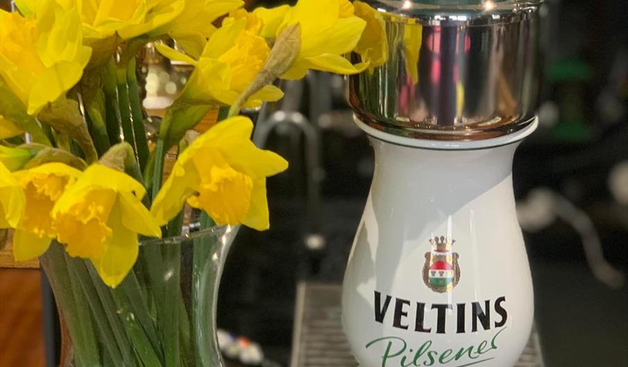 Interior of The Ship Inn. A glass vase filled with bright yellow daffodils is placed next to a white and chrome Veltins Pilsener beer tap on a bar cou