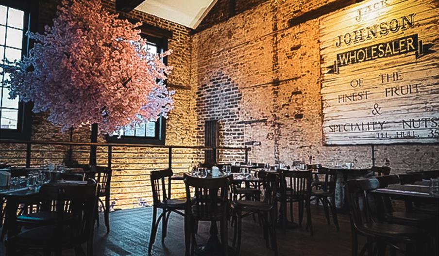 Restaurant with high ceilings, wooded floors, exposed brick walls, wooden table and chairs  with a faux, pink cherry tree in the corner.