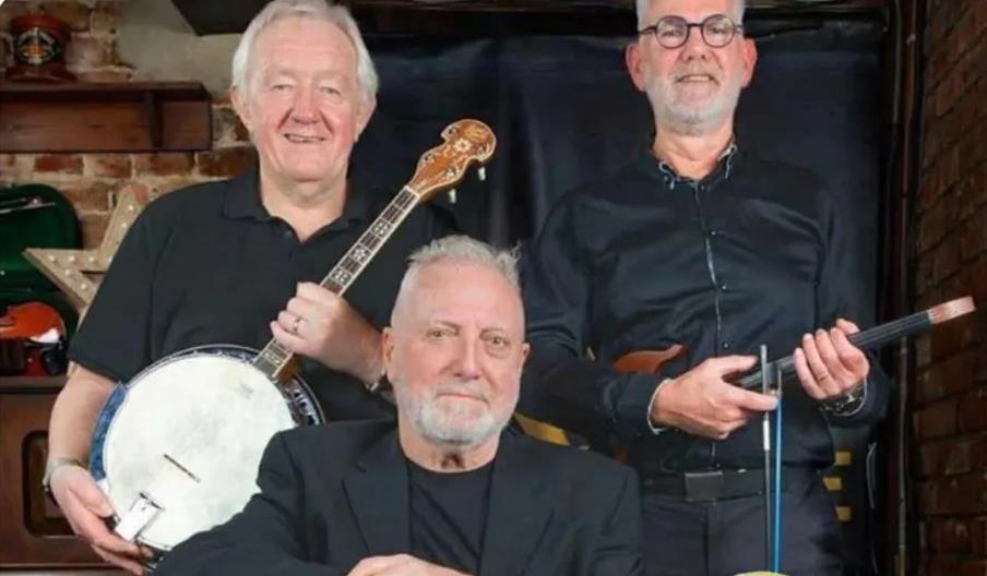Three older men pose together indoors; one holds a banjo, another holds a violin, and the third sits in front, suggesting they are a musical group.