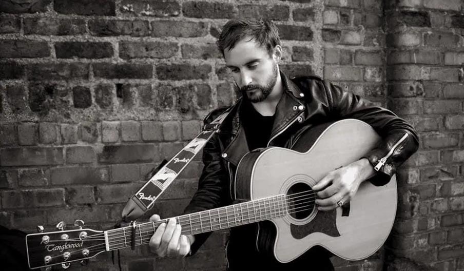 A man wearing a leather jacket plays an acoustic guitar in front of a brick wall, looking down at the instrument.