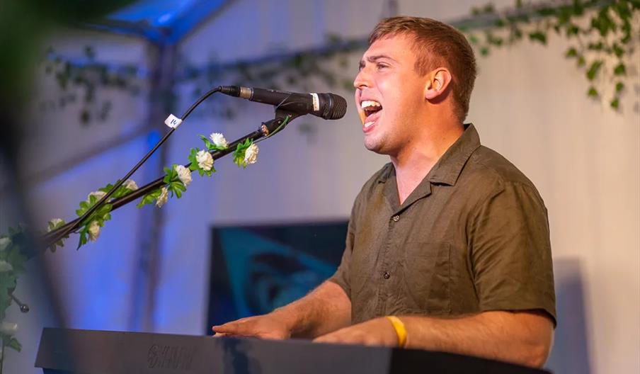 A man passionately sings into a microphone while playing a keyboard on stage, surrounded by greenery and floral decorations.