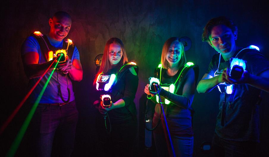 Four people in the dark wearing multicoloured light up armour and pointing laser rifles at the camera.