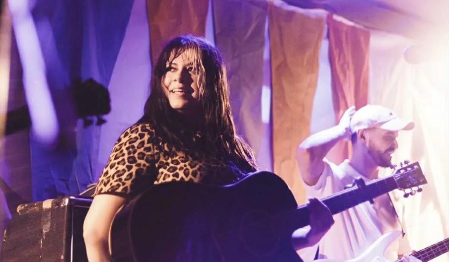 A woman with long dark hair wearing a leopard print shirt plays an acoustic guitar on stage, while a man in a white shirt and cap stands behind her pl