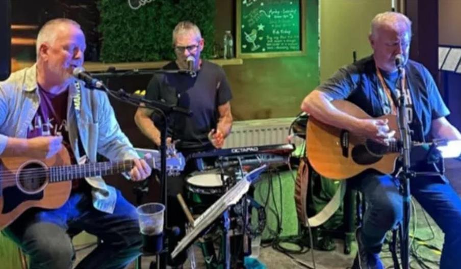 Three middle-aged men perform live music indoors; two play acoustic guitars and sing into microphones, while the third plays a keyboard and drum, also