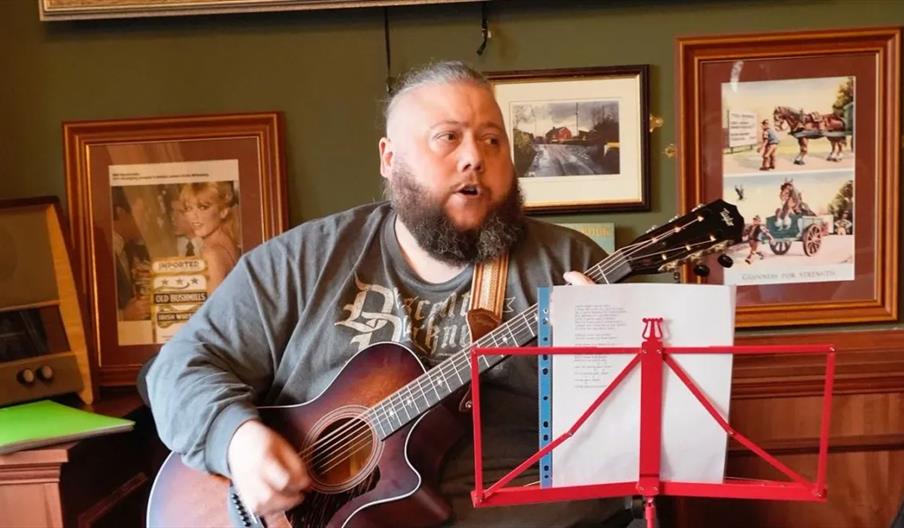 A bearded man is playing an acoustic guitar and singing, seated indoors with framed pictures on the wall behind him. A music stand with sheet music is