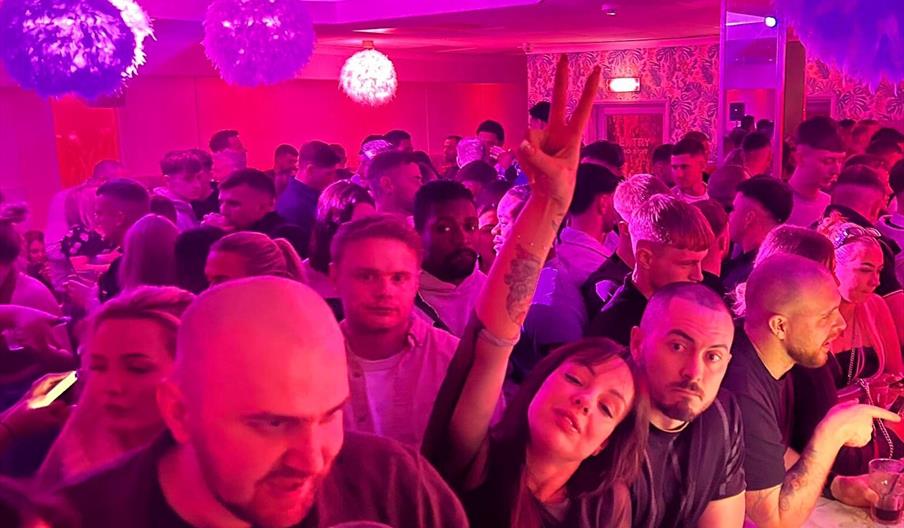 Interior of Pink Parrot. A crowded nightclub scene with people gathered closely together under purple and pink lighting, some holding drinks, and one