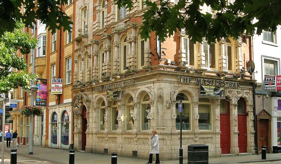 A person walks past an ornate, historic building housing "The Masters Bar" on a city street, with red doors, arched windows, and intricate stonework u